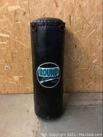 Full view of the black Round One punching bag standing upright against a wooden panel wall, showing overall shape and logo.