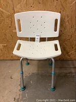 Front view of shower chair showing plastic seat and backrest with handles, aluminum legs, and rubber feet on concrete floor against wooden wall.
