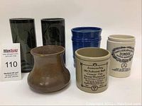 Front view of the six stoneware items including two dark green glazed vases, one brown bulbous vase, and three cylindrical marmalade jars in blue, white, and beige.