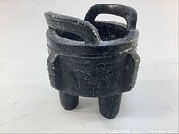 Side view of the black soap stone footed dish showing carved faces and two curved handles above the rim, standing on three legs.