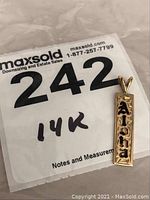 Photo of the gold pendant front side showing 'Aina' in black enamel on textured gold surface attached to sale tag with number 242 and '14K' written on it.