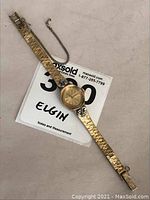 Full view of ladies Elgin wristwatch laid flat showing gold-tone patterned bracelet and safety chain attached near clasp.