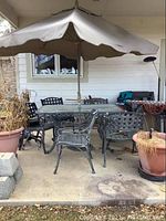 Outdoor metal table with glass top and beige umbrella in center, surrounded by six matching metal chairs on concrete floor.