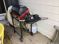 Full view of the Mecco electric outdoor grill on black stand with wheels, with accessories and cover beside it on the patio floor.