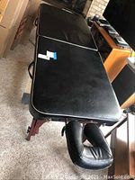 Full view of black folding massage table with attached face cradle in upright position on carpet.
