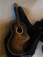 Full view of the Applause acoustic guitar in its soft case on carpeted floor, showing black glossy finish and wooden bridge.