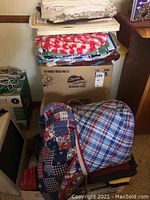 Stack of assorted folded table linens including various placemats and napkins, with a folded 4th of July themed patchwork picnic quilt in red, white, and blue colors.