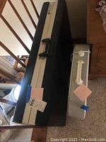 Folded Battle Creek portable massage table in black and beige with a black handle placed on a floor next to other items.