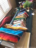 Photo showing colorful woven sarape blanket atop folded quilt with books and decorative items along the table.