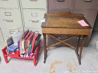 Photo showing the children's wooden antique desk with a red basket of art and educational supplies next to it, placed on a floor with filing cabinets in the background.