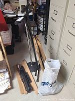 Photo showing metal microphone stands, a white bag labeled 'Microphone,' and some cardboard boxes in a basement setting.