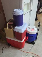 Lot of five coolers stacked including large red wheeled cooler and blue wheeled sports cooler.