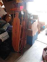 Two vintage Flexible Flyer snow sleds leaning against a wall surrounded by packed boxes and miscellaneous items.