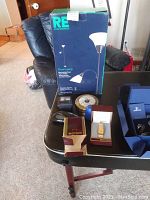 Items on black table including Citizen watch box with gold-tone watch, barometer in gold trim, two travel clocks and boxed REI floor lamp.