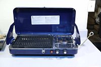 Full view of the blue Woods propane camp stove with lid open showing grill grate on left and burner on right.