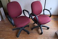 Photo showing two red fabric office chairs with black plastic armrests and bases with wheels, positioned side by side on a wooden floor.