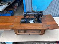 Full view of vintage Singer sewing machine mounted in a wooden table base without legs, showing fold-out table extensions on both sides.