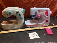 Two mini toy sewing machines, one Perfect Stitch cream colored, one white Singer with pink accents, ruler and pink foot pedal shown