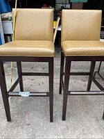 Pair of tall counter bar chairs with cracked tan vinyl seats and dark wood frames showing chips and scratches.
