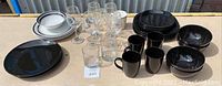 Overview of the dishware and glassware lot showing white and black plates, black bowls, black mugs, and clear glassware with stems and tumblers.