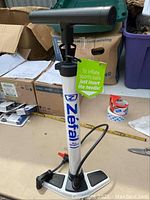 Full view of the white Zefal bicycle pump with blue branding and black handle and base, showing the tag indicating it can inflate sports balls by inserting the needle.