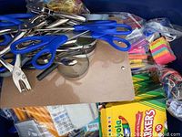 Photo showing blue handled scissors, metal hole punches, colored markers, sticky notes, clipboards, pencils, and a white calculator in a plastic tub.