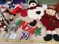 Photo of the lot showing plush holiday figures including Santa Claus, snowman, and knit Santa figure alongside decorative signs spelling Noel and Joy and green Christmas tree ornaments.