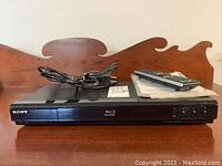 Front view of the Sony Blu-ray DVD player on a wooden surface showing the device, power cord, remote control, and manual.
