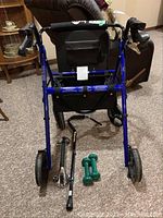 Walker viewed from the front showing four wheels, padded seat, and accessories on the floor (cane, dumbbells, bath grab bar).