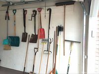 Garage wall overview showing brooms, dustpan, snow shovel, square shovels, hoe, cultivator and rakes hung on hooks