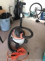 Wide view of Rigid wet/dry shop vacuum sitting on the floor with attached black hose, orange extension cord coiled on floor, and garage background with bike and garbage bin.
