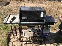 Full view of black Weber propane gas grill with side tray holding grill tools, propane tank attached below, grill standing on stone patio