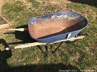 Side view showing the full length of the wheelbarrow with blue metal tub, wooden handles, and rubber tire on grass.