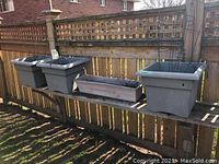 Four grey resin garden planters lined on wooden shelf outdoors with some containing tomato cages and minimal soil.