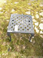 Square cast iron patio table photographed outdoors on mossy stone ground, showing lattice top and legs from multiple angles.