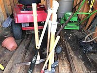 Group of axes, sledgehammer, pick axe, and baseball bat in a wooden shed on the floor with other garden equipment visible.
