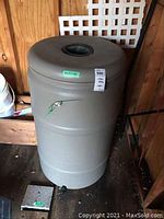 Beige cylindrical plastic rain barrel with spigot seen indoors on wooden floor.