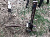 Full view of the manual high-lift car jack resting on grassy ground, showing rust and wear on metal parts.