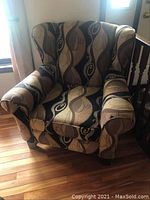 Front and angled front view of an upholstered armchair with contemporary swirling pattern upholstery in beige, tan and dark brown colors, placed on wood flooring.