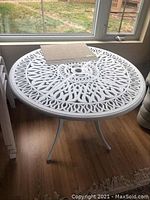 Front angle view of white round metal patio table with detailed cutout design and umbrella hole, indoor setting.