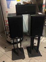 Two black Energy cabinet speakers on stands with an Advent center speaker and speaker wire on top, photographed in basement near water heater.