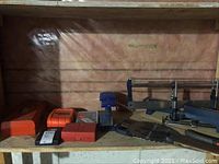 Wide view of shelf showing all items including orange and red drill bit cases, blue case, saw blades, frame saw with miter box, and hand saws.