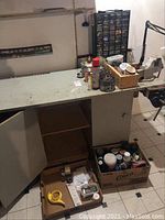 Workshop table corner with open cabinet below, containing cardboard box with tape measures and other small hardware, a box with spray cans and containers.