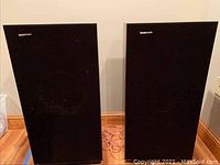 Two black rectangular Boston Acoustics speakers side by side on floor, front view with fabric grills.