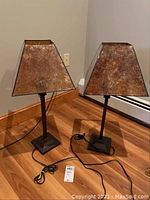 Pair of two lamps side by side showing metal bases and trapezoidal shades with mottled brown color on hardwood floor in corner of room.