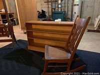 Wooden bookcase with three shelves alongside wooden rocking chair with rattan seat, shown on a black surface in basement setting.