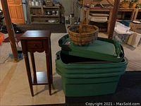 Photo showing four large green storage containers stacked with lids, a round straw basket placed on top, and a small wooden side table with drawer and lower shelf nearby.
