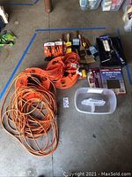 Photo showing electrical cords, paint brushes with wooden handles, cans of painting supplies, paint trays, and sanding sheets on concrete floor.
