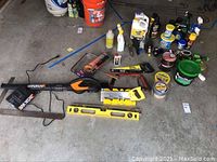 Full view of assorted tools and yard supplies laid on concrete floor including leaf blower, hand saw, leveler, duct tape and spray bottles