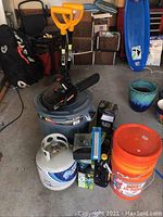 Overview of lot showing chainsaw on top of trash bin, snow shovels behind, propane tank and torches in original packaging in front, and 3 orange buckets on the side.